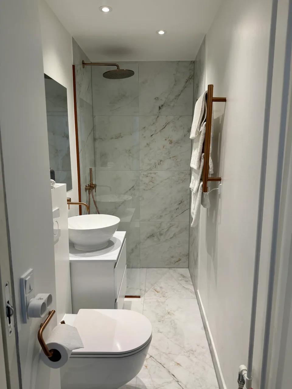 Nyt badeværelse med marmorfliser fra gulv til loft, væghængt toilet, håndvask på skab og walk-in-bruser bag glas.