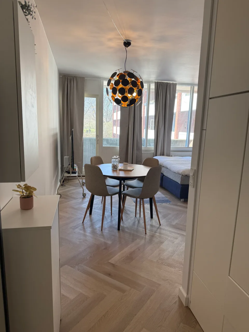Kig gennem lejligheden efter renovering: spisebord under en messingfarvet pendel, sildebensgulv og soveplads bag ved de lyse gardiner.