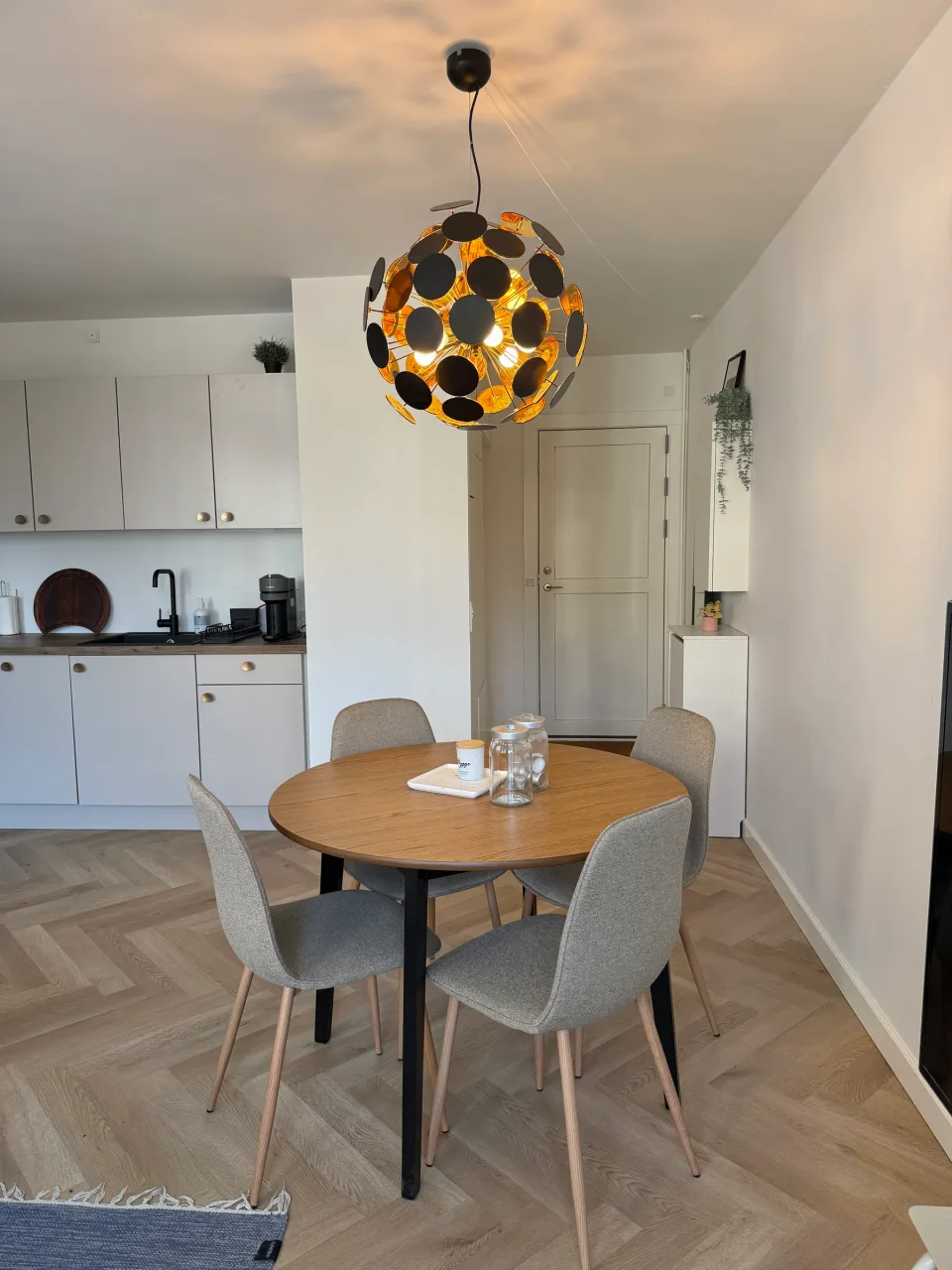 Alrum efter renovering med sildebensgulv i lys eg, rundt spisebord i egetræ, grå polstrede stole og et nyt lyst køkken langs væggen.